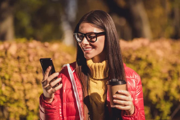 긍정적 인 소녀 블로거의 사진 휴식 도시 가을 공원 음료 takeaway 커피 라떼 음료 mug 사용 스마트폰 스마트폰처럼 보이는 소셜 미디어 포스팅빨간 옥시 재킷 — 스톡 사진