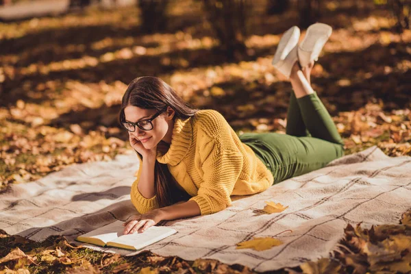 Полноразмерная фотография позитивной девочки, отдыхающей в загородном осеннем лесу, лежащей в обшарпанном одеяле с книгой для чтения в желтом цвете. — стоковое фото
