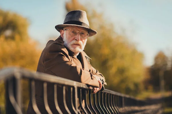 Profile photo of retired old white hair grandparent man street central park walk lean metal fence bridge near river serious facial expression wear vision spectacles autumn jacket hat outside