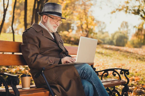 Perfil lado de la foto del viejo hombre positivo tienen en línea familia portátil comunicación turismo tipo de correo electrónico sentarse banco en otoño naturaleza follaje árboles centro de la ciudad parque desgaste abrigo gorra sombrero — Foto de Stock