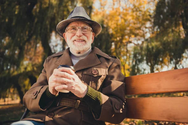 Foto de hombre viejo inspirado sentarse banco bebida celebrar comida para llevar taza de café sueño soleado amarillo colorido sol tiempo otoño parque naturaleza descanso relajarse usar gorra chaqueta de la cabeza — Foto de Stock