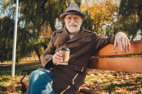 Foto de hombre viejo positivo descanso relajarse octubre otoño fin de semana parque pausa sentarse banco disfrutar aromático para llevar taza de café desgaste gorra chaqueta de la cabeza — Foto de Stock