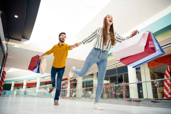 Vista de ángulo bajo de longitud completa foto de loco emocionado dos cónyuges personas ver increíble off-sales correr prisa rápida celebrar muchos paquetes de desgaste de rayas vaqueros camisa amarilla en el centro comercial centro comercial — Foto de Stock