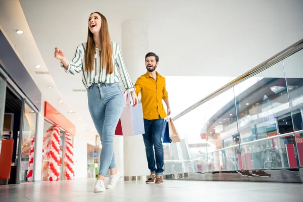 Vista de ángulo bajo de longitud completa foto de la mujer enérgica ver increíbles 50 ventas ir a pie prisa su marido seguir mantenga muchos paquetes de desgaste de rayas vaqueros camisa amarilla en el centro comercial centro comercial — Foto de Stock