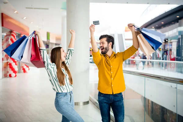 Foto de duas pessoas alegre bonita senhora bonito cara casal desfrutar de tempo livre segurar muitos sacos andar shopping center levantar as mãos usar cartão de crédito descontos desgaste casual jeans camisa roupa dentro de casa — Fotografia de Stock