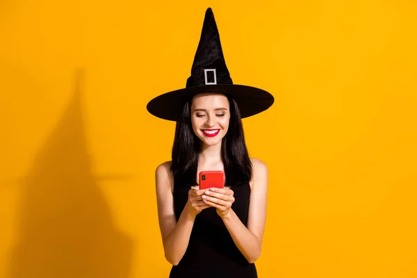 可愛い若い魔術師の女性の写真は、電話の笑みを浮かべて見て画面を保持友人の最近の記事を読むお気に入りの着用黒ウィザードのヘッドウェアドレス孤立明るい黄色の色の背景 — ストック写真