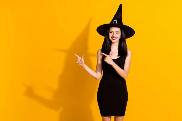 かわいい魅力的な若い魔術師の女性の直接指の空のスペース光沢のある笑顔の写真は、魔女の服黒のヘッドウェアの短いミニドレスを提案孤立明るい黄色の色の背景 — ストック写真