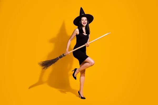 フルボディサイズの写真のかなり魅力的な若い魔術師女性笑顔ホールドbroomstickふり空の飛行は黒の帽子を着用高ヒールドレス孤立明るい黄色の色の背景 — ストック写真