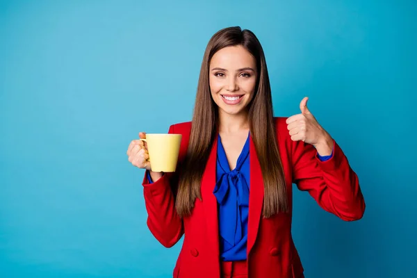 Foto de atractiva dama de negocios morena sosteniendo taza de café caliente por la mañana levantar el pulgar hacia arriba aconsejando ropa de increíble calidad elegante traje rojo con camisa aislada de color azul de fondo — Foto de Stock