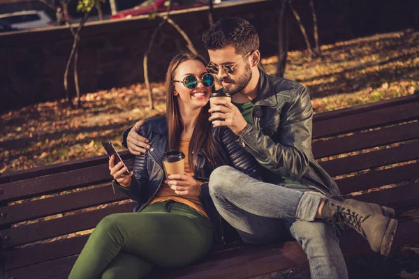 Фото двох студентів сідають на лавку в жовтому міському парку осінь використовувати смартфон пити каву виймання напій кухоль — стокове фото