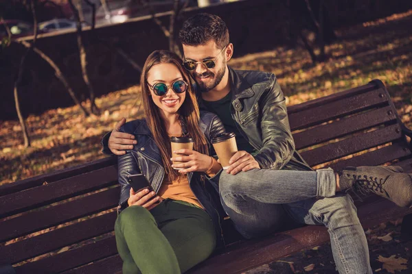 Foto de positivo duas pessoas casal sentar banco no outono setembro parque da cidade uso smartphone bebida takeout café caneca bebida — Fotografia de Stock