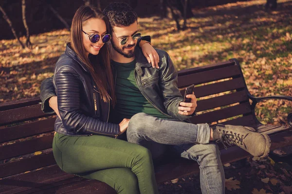 Фото позитивних двох студентів сідають на лавку в міському осінньому парку, читають пости в соціальних мережах, одягнені в сонячну куртку — стокове фото