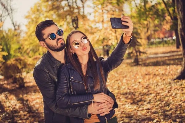 Фото двох студентів, які обіймають дівчину, роблять селфі на мобільний телефон, відправляють повітряний поцілунок восени вересень міський парк одягають повсякденну куртку — стокове фото