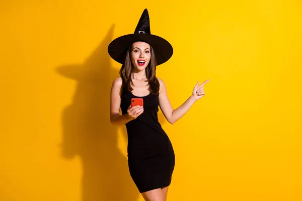 Fotoğraf için heyecanlı cadı kadın akıllı telefon noktası işaret parmağı telif alanı gizemli büyüleyici reklamlar siyah şapka etek giyer parlak parlak arkaplan — Stok fotoğraf