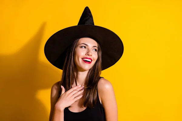 Foto de encantadora jovem senhora sorrindo vermelho brilhante batom coquete olhar para cima espaço vazio mão peito queda evento desgaste preto bruxa traje cone headwear isolado cor amarela fundo — Fotografia de Stock