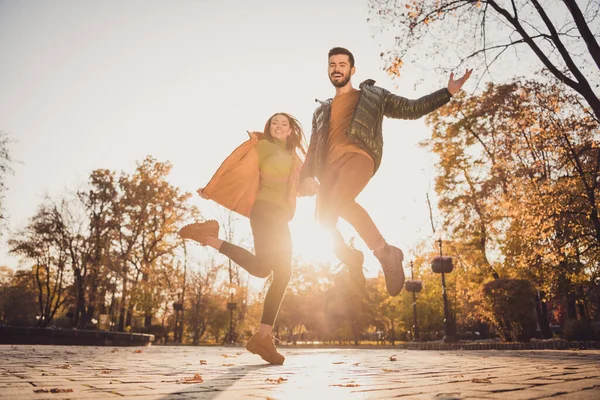 Low angle view full length photo of cheerful two young people girl guy hold hand jump in autumn october town park — 스톡 사진
