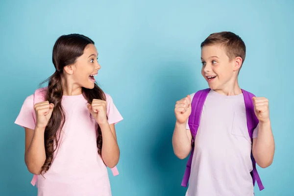 Foto extático emocionado dos niños de la escuela niña niño impresionado recibir un resultado de prueba de nivel levantar los puños gritar sí se miran desgaste mochila bolso camiseta aislado color azul fondo —  Fotos de Stock