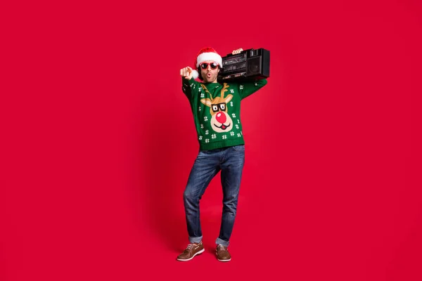Full length body size view of attractive funny cool hipster guy wearing Santa hat carrying boombox having fun pointing choose choice you isolated bright vivid shine vibrant red color background — Zdjęcie stockowe
