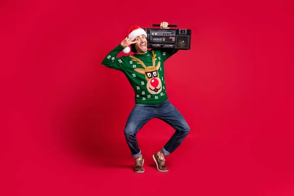 Взгляд в полный рост на привлекательного, безумного, жизнерадостного веселого парня в шляпе Санта-Клауса, танцующего, неся в руках коробку с V-образным знаком на ярко светящемся красном фоне — стоковое фото