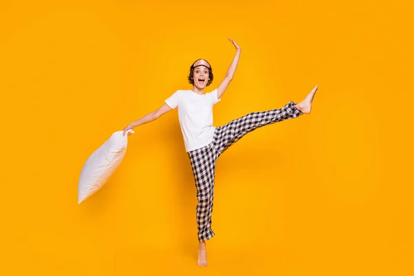 Full body photo of cheerful lady hold big pillow wake up playful mood dancing energetic raise leg up wear mask white t-shirt plaid pajama pants barefoot isolated yellow color background