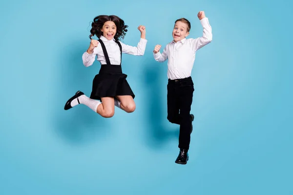 Фотографія двох протилежної маленької дівчинки хлопчик школярі брат сестра однокласники стрибають кулаки в останній шкільний день одягнені білі сорочки чорні штани сукні ізольовані синій колір фону — стокове фото