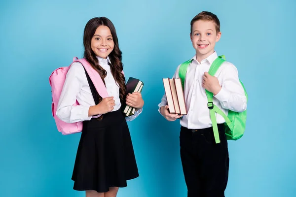 Фото двух маленьких девочек школьник брат сестра одноклассники держать книги ходить библиотека учебного года заканчивая носить рюкзак белая рубашка черные брюки платье изолированный синий цвет фона — стоковое фото