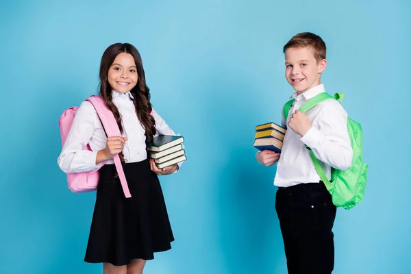 Foto de dos niños pequeños colegiales hermanos compañeros de clase sostienen libros de pila biblioteca año de estudio que comienza a usar mochila camisa blanca pantalones negros vestido aislado color azul fondo —  Fotos de Stock