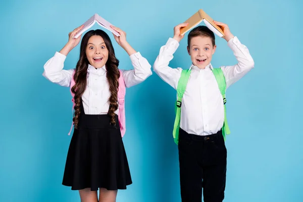 Фото двух маленьких девочек мальчик школьники брат сестра одноклассники держать учебник над головой весело школьный перерыв носить мешок белая рубашка черные брюки платье изолированный синий цвет фона — стоковое фото