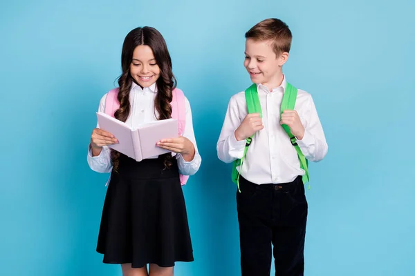 İki küçük kız öğrencinin fotoğrafı okul sonrası en iyi arkadaşları ödevlerini hazırlar ders kitabı okurlar sırt çantası giyerler, beyaz gömlek giyerler, siyah pantolon giyerler izole edilmiş mavi arka plan. — Stok fotoğraf