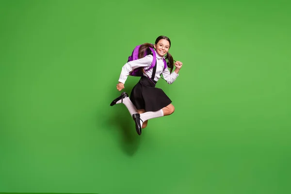 Foto portrét brunetky školačka s copánky běží skákání nahoru s fialovým batohem izolované na živém zeleném pozadí — Stock fotografie