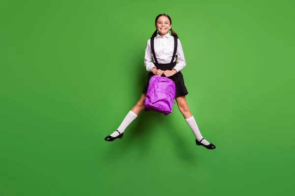 Retrato fotográfico de una colegiala feliz saltando con las piernas extendidas sosteniendo la mochila púrpura aislada sobre un fondo verde vivo —  Fotos de Stock