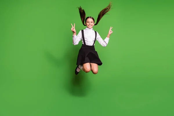 Foto retrato de colegial morena com tranças pulando segurando dois v-sinais isolados no fundo de cor verde vívido — Fotografia de Stock