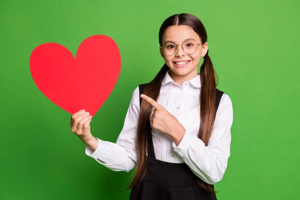 Foto de muito adorável pequena senhora estudante na frente de quadro-negro direto dedo mostrar papel vermelho coração figura dia dos namorados promo desgaste branco camisa óculos isolado cor verde fundo — Fotografia de Stock