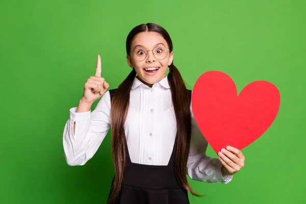 Foto retrato de colegial morena segurando grande cartão postal coração vermelho apontando dedo para cima isolado no fundo de cor verde vívido — Fotografia de Stock