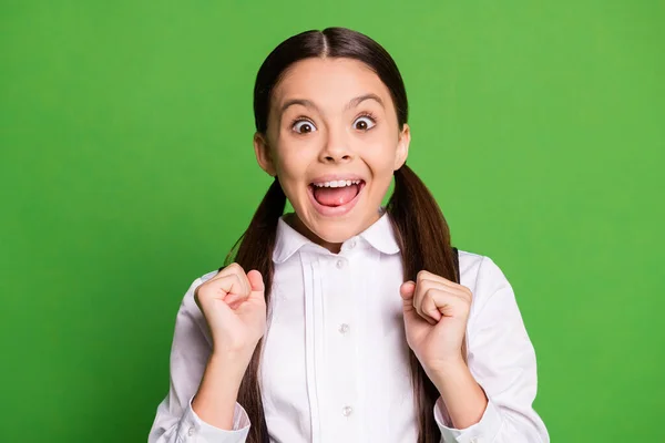 Foto av söt söt liten dam hästsvans frisyr skolbarn upphetsad öppen mun håll knytnävar vinna biljetter bio klasskamrater bjuda in bära uniform vit skjorta isolerad grön färg bakgrund — Stockfoto