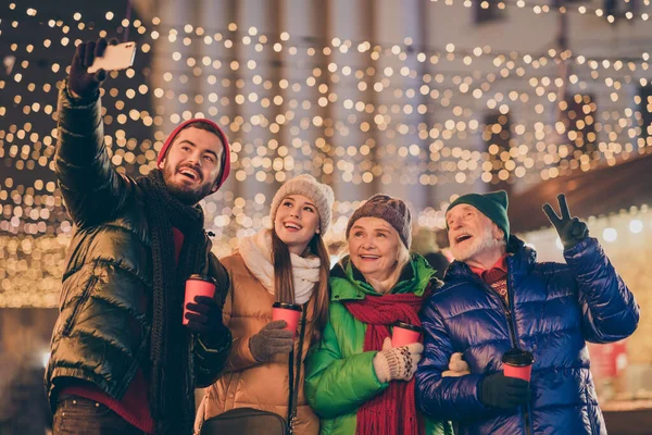 Фото всей семьи четыре человека x-mas сбор держать кружку сделать селфи телефон шоу V-знак носить верхнюю одежду шляпу пальто перчатка многопоколения ночной улице освещения на открытом воздухе — стоковое фото