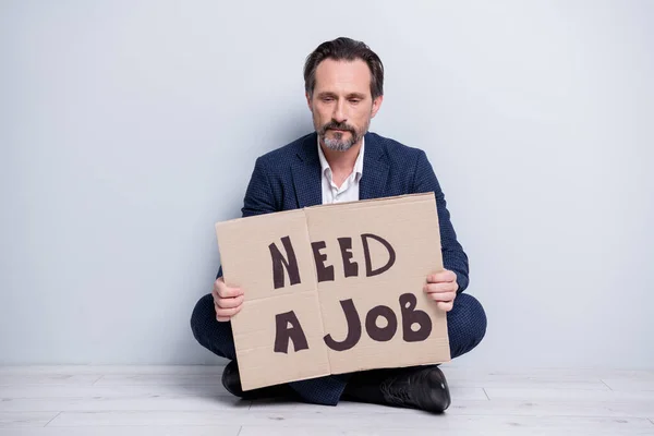 Full length photo of unhappy worker mature guy financial crisis lost work hold carton placard banner search work place moody sitting floor wear blue suit shoes isolated grey background — Stock fotografie