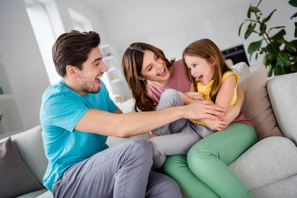 Foto de familia alegre positiva tres personas mamá papá jugar pequeña niña cosquillas risa sentarse sofá en casa en el interior — Foto de Stock