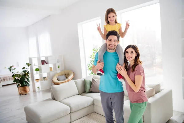 Foto de positivo tres personas mamá papá llevar hombros niña pequeña niño hacer v-signo en casa en el interior —  Fotos de Stock