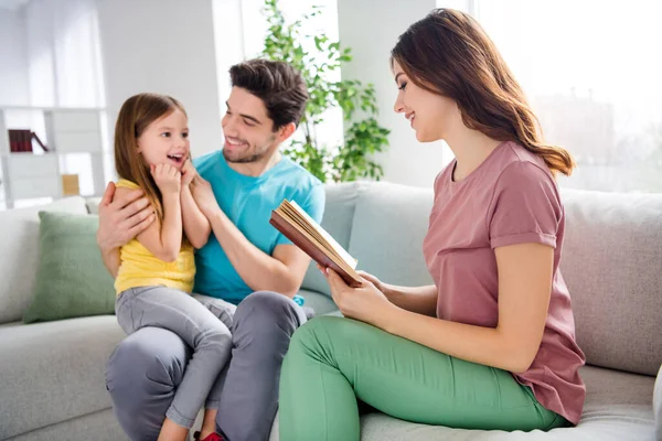 Foto de tres personas emocionadas niña pequeña sentarse padre rodillas sofá escuchar a su madre leer libro cuentacuentos en casa en el interior — Foto de Stock