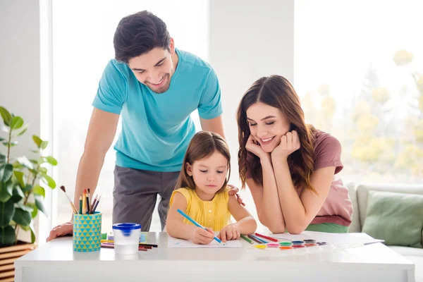 Foto de apoyo positivo confianza tres personas mamá papá se siente orgullosa pequeña hija sentarse mesa dibujar color lápiz papel foto en casa en interiores — Foto de Stock