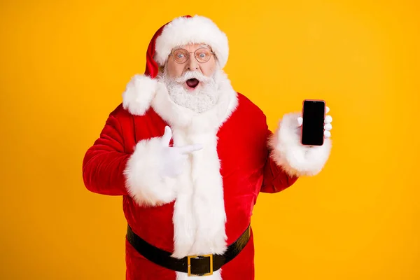 写真脂肪白い灰色のひげの毛サンタクラスポイント指のスマートフォンは最高のx-マスシーズンショッピングクリスマスギフトの選択は、赤の帽子のヘッドウェアを身に着けています孤立した黄色の背景 — ストック写真