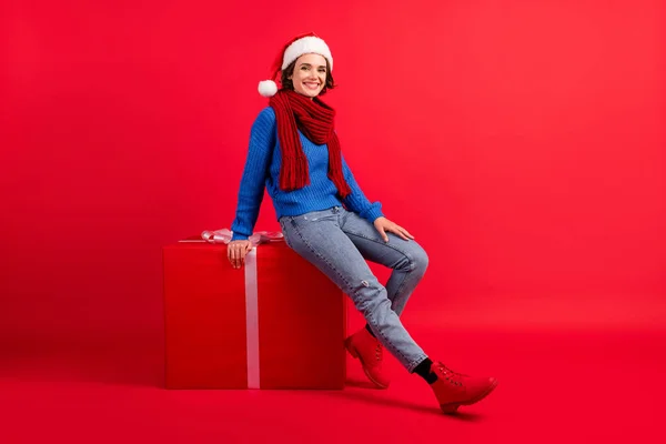 Foto de longitud completa chica positiva en santa claus headwear sentarse caja de regalo grande disfrutar de x-mas navidad nuevo año fiesta evento desgaste estilo jersey denim botas aisladas brillo brillante color fondo — Foto de Stock