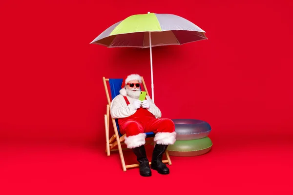 Портрет його гарний красивий бородатий жир Санта сидить у кріслі, використовуючи сервіс пристрою роумінгу блог холодний тропічний тур курортний тур сонячний день ізольований яскравий яскравий блиск яскравий червоний колір фону — стокове фото