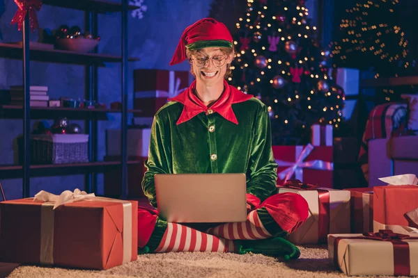 Foto de comprimento total de elfo sentar-se tipo de saudação no laptop usar óculos verdes cap em casa dentro de casa com luzes de decoração noite x-mas — Fotografia de Stock