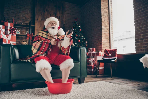 Full length photo white grey hair beard santa claus sit couch relax x-mas gift north delivery watch comedy switch channel wear red cap headwear glasses in house indoors christmas eve tree