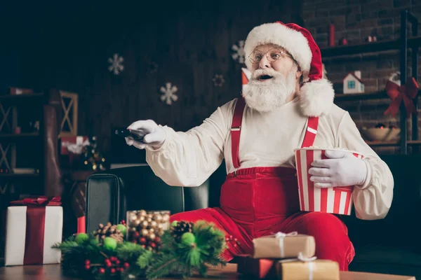 令人惊讶的灰胡子桑塔爪坐在沙发上看圣诞片吃爆米花盒开关频道头戴红帽子在屋里的诺瓦装饰品 — 图库照片