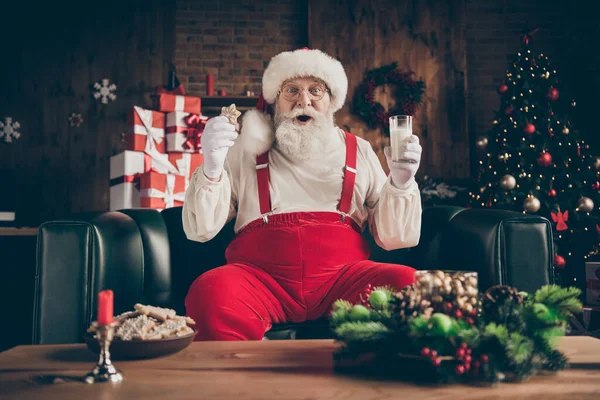 Choqué barbe grise santa claus assis confortable canapé regarder x-Noël film de cinéma impressionné boisson verre de lait manger pain d'épice cookie porter casquette rouge casque dans la maison ornement noel intérieur — Photo