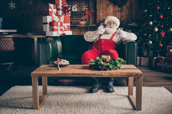 Полная длина фото веселой седой бороды Санта Клаус сидеть комфортный диван наслаждаться смотреть рождественский фильм сказочный напиток молоко едят печенье носить головной убор красный колпак в Ноэль пришествие оранжерея дом в помещении — стоковое фото