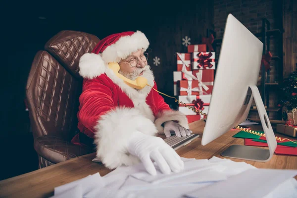 Profilseite Foto von grauen bärtigen Weihnachtsmann sitzen Tisch überprüfen Computer E-Mail-Anruf Telefon zustimmen x-mas Weihnachtsgeschenk Geschenk Lieferzeit tragen rote Mütze Kopfbedeckung im Haus drinnen — Stockfoto